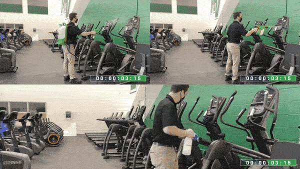 A person wearing a mask and gloves cleans gym equipment with a spray bottle and cloth, wiping down elliptical machines in a well-lit fitness center.